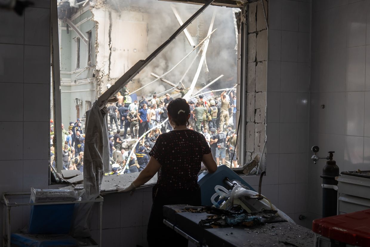 A healthcare worker looks through a window of a damaged operating room at people clear rubble at the building of one of the largest children’s hospitals of Ukraine, Okhmatdyt