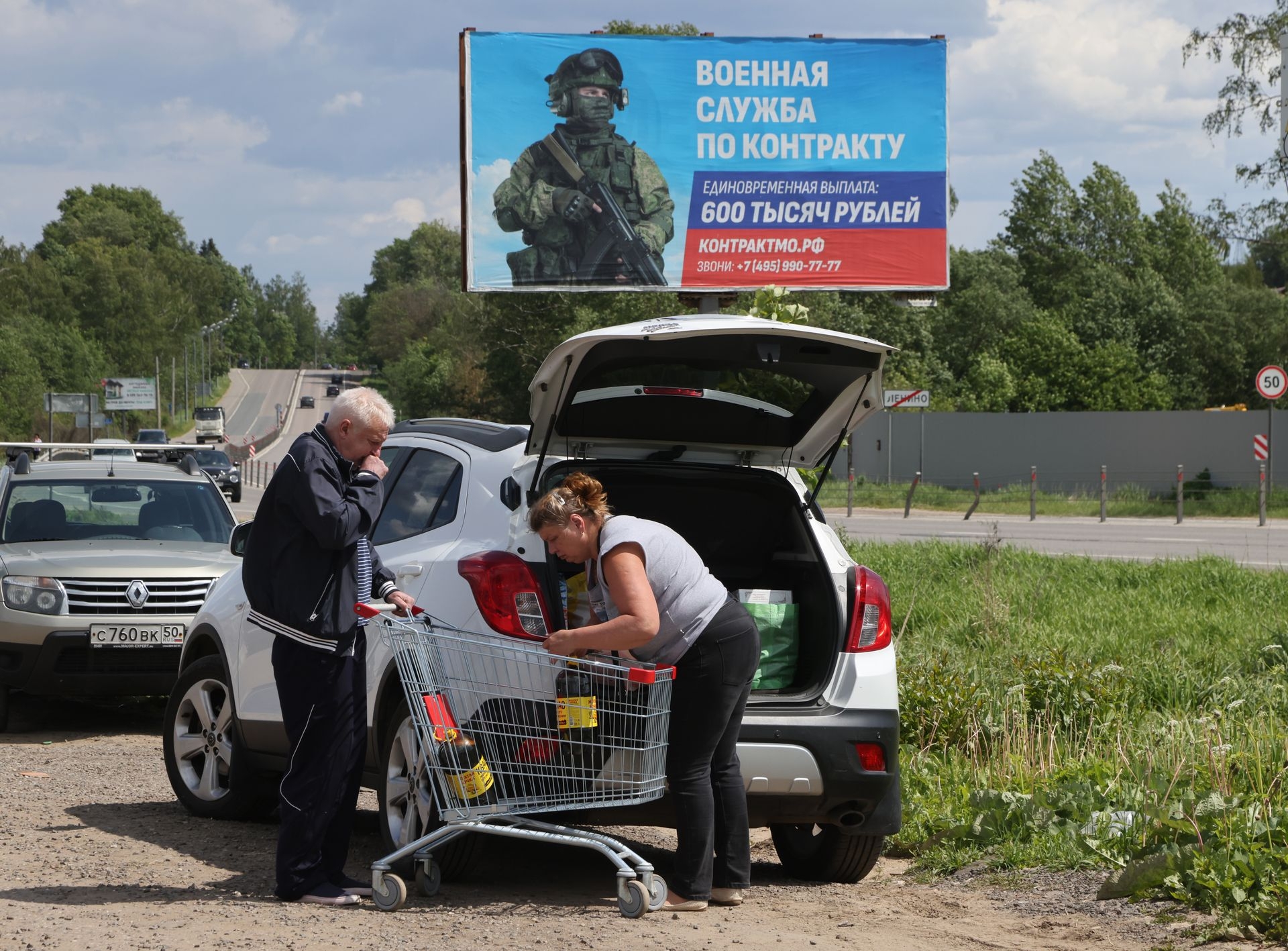 An elderly couple loads the trunk of their car with purchases near a supermarket as a military propaganda billboard stands in the background in Lenino, Russia, on June 7, 2023