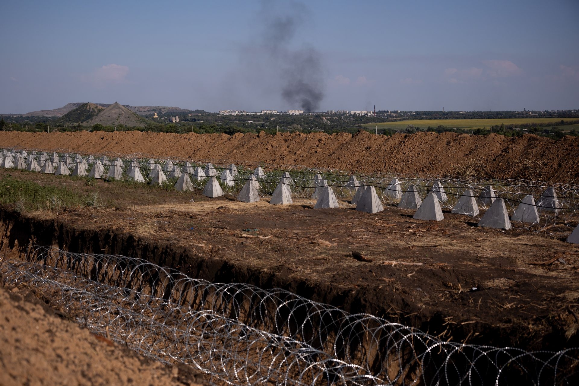 Fortifications, including dragon’s teeth and barbed wire, are seen in front of smoke rising over Dobropillia after the detonation of an aerial bomb in Rodynske, Donetsk Oblast, Ukraine, on July 16, 2025.