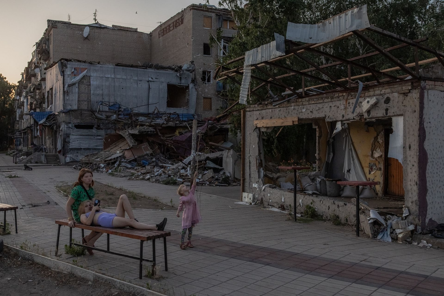 A child plays as women rest on a bench in front of residential buildings destroyed by shelling in Kostyantynivka, Donetsk Oblast, Ukraine, on June 22, 2024, amid the Russian invasion.