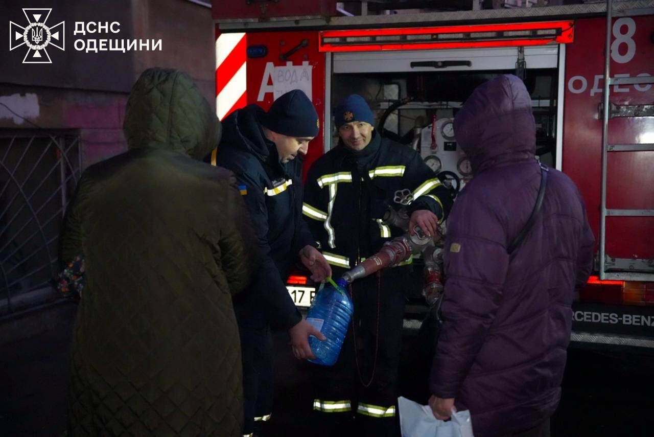 Two emergency responders dispense water from a red firetruck for local residents standing aside