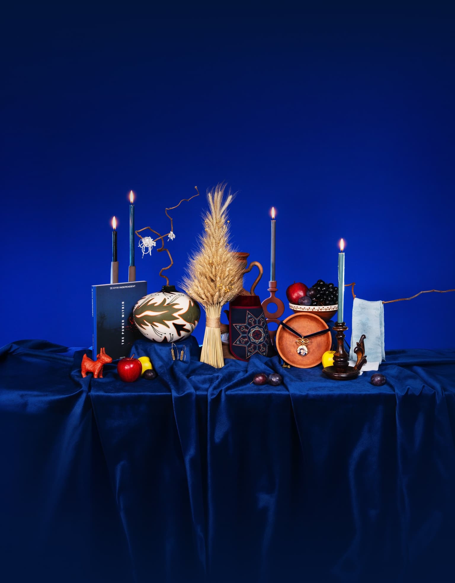 A still life on blue velvet: lit candles, a wheat didukh, a painted ceramic vase, a clay jug, the Kyiv Independent book The Power Within, a clay plate with a pendant, and fruit