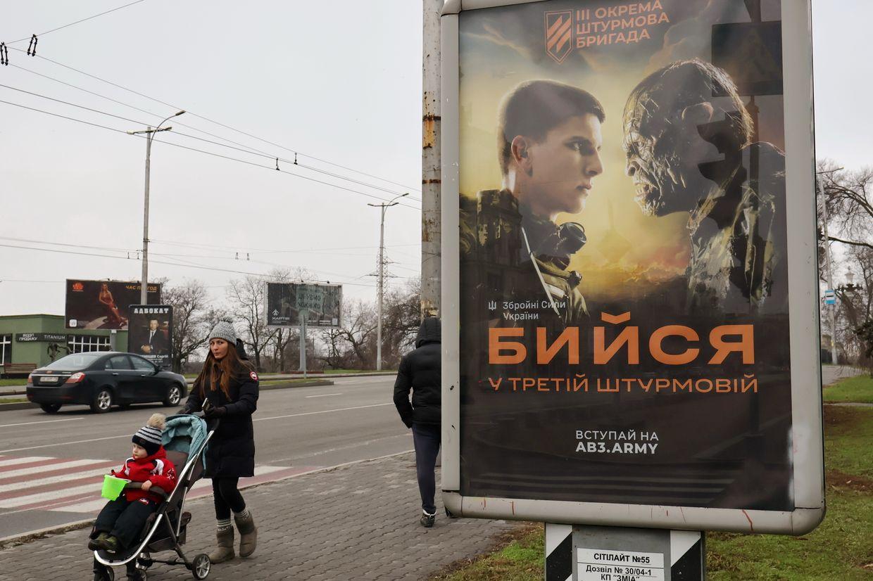 A woman with a child in a baby carriage walks past the recruitment poster for the Ukrainian armed forces