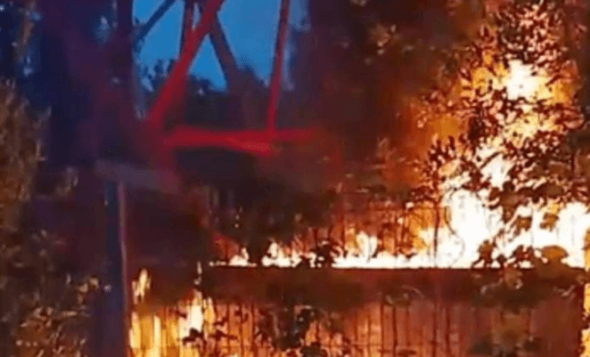 A fire burns under a metal structure, around a fence, and bushes following an alleged strike.