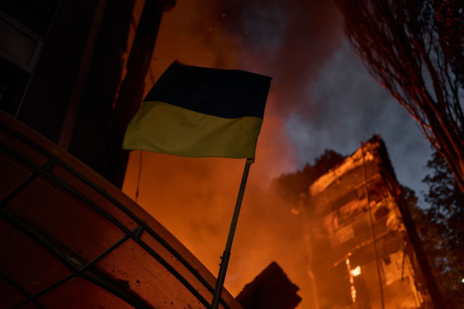 A Ukrainian flag flies in front of a fire following a mass Russian attack on the capital in Kyiv, Ukraine, on July 4, 2025. 