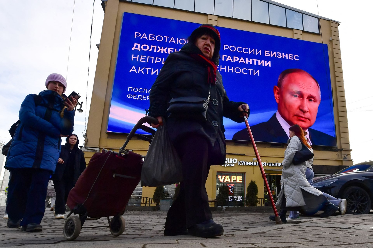 People walk past a digital screen displaying Vladimir Putin and a quote from his address to the Federal Assembly in Saint Petersburg, Russia, on March 13, 2024.