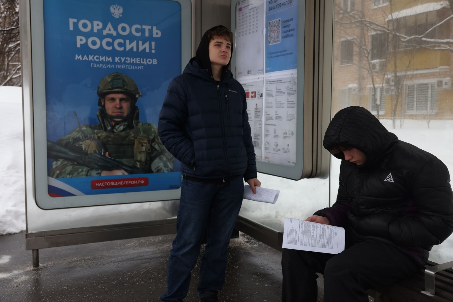 Russian students wait at a bus stop next to a military propaganda poster depicting a participant of the invasion of Ukraine, in Moscow, Russia, on Feb. 25, 2026.