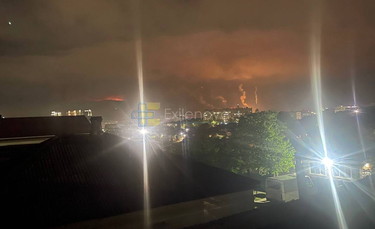 Smoke rises from an orange glow in the distance behind houses, trees, and a building.