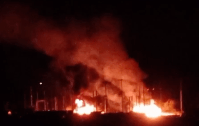 Heavy smoke rises from a burning fire within a fenced area under a dark night sky