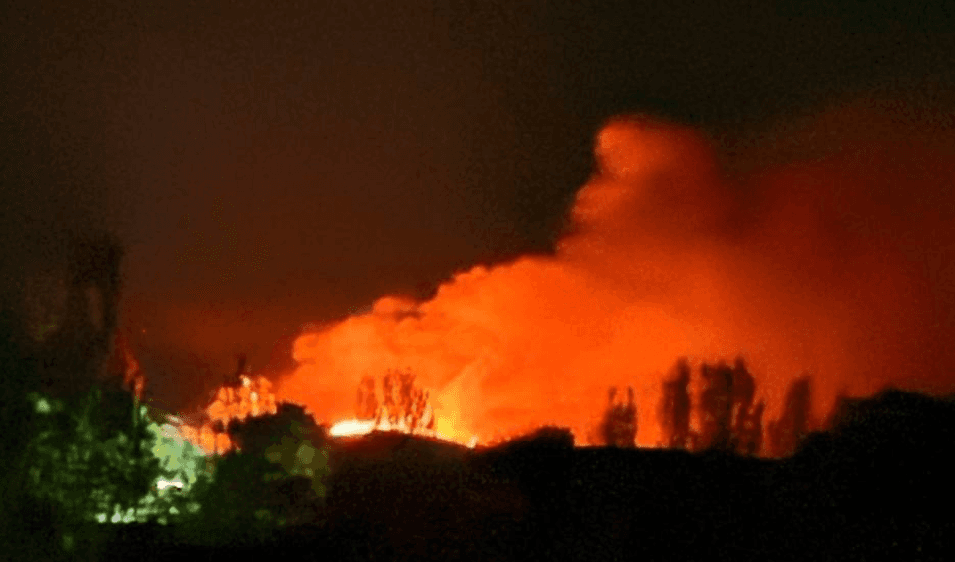A large fire burns behind a treeline under a night sky