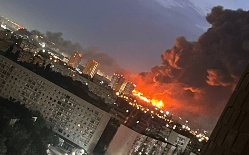 A massive fire burns in the distance behind several blocks of mid-rise residential buildings