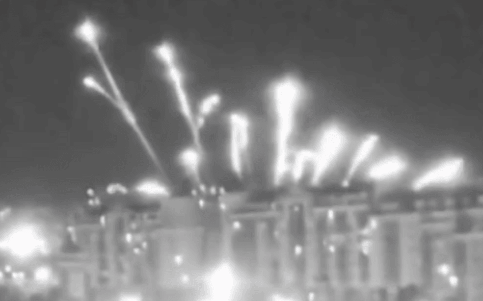 Several small explosions in the sky over a a block of buildings in a screenshot of black and white footage
