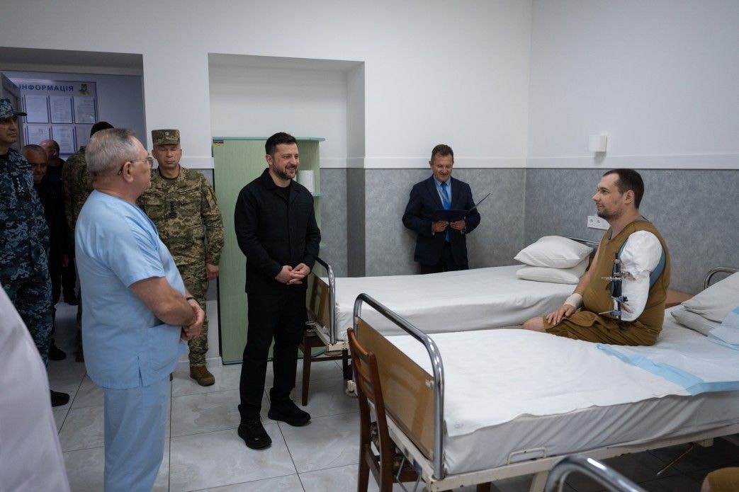 Presisdent Volodymyr Zelensky, wearing all black stands by the bedside of a wounded Ukrainian soldier in a medical facility.