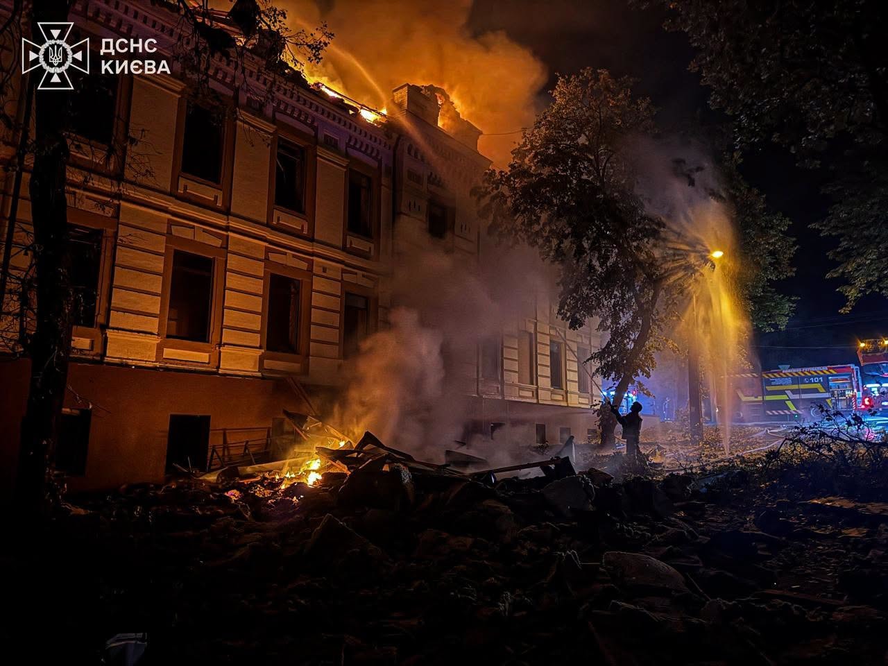 A 2-story historic-style building burning with its windows shattered has 1 firefigther pointing a stream of water at its roof