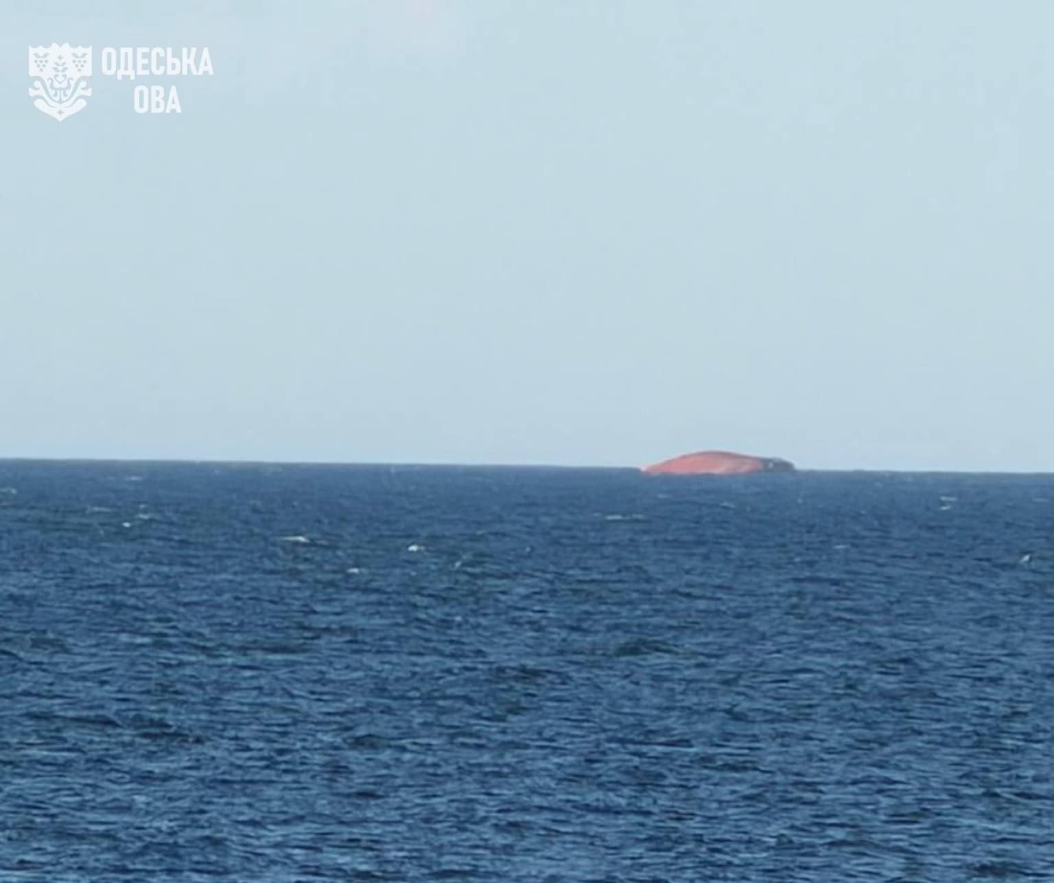 A capsized cargo vessel with an orange/red belly and dark hull elsewhere floats on a deep blue sea